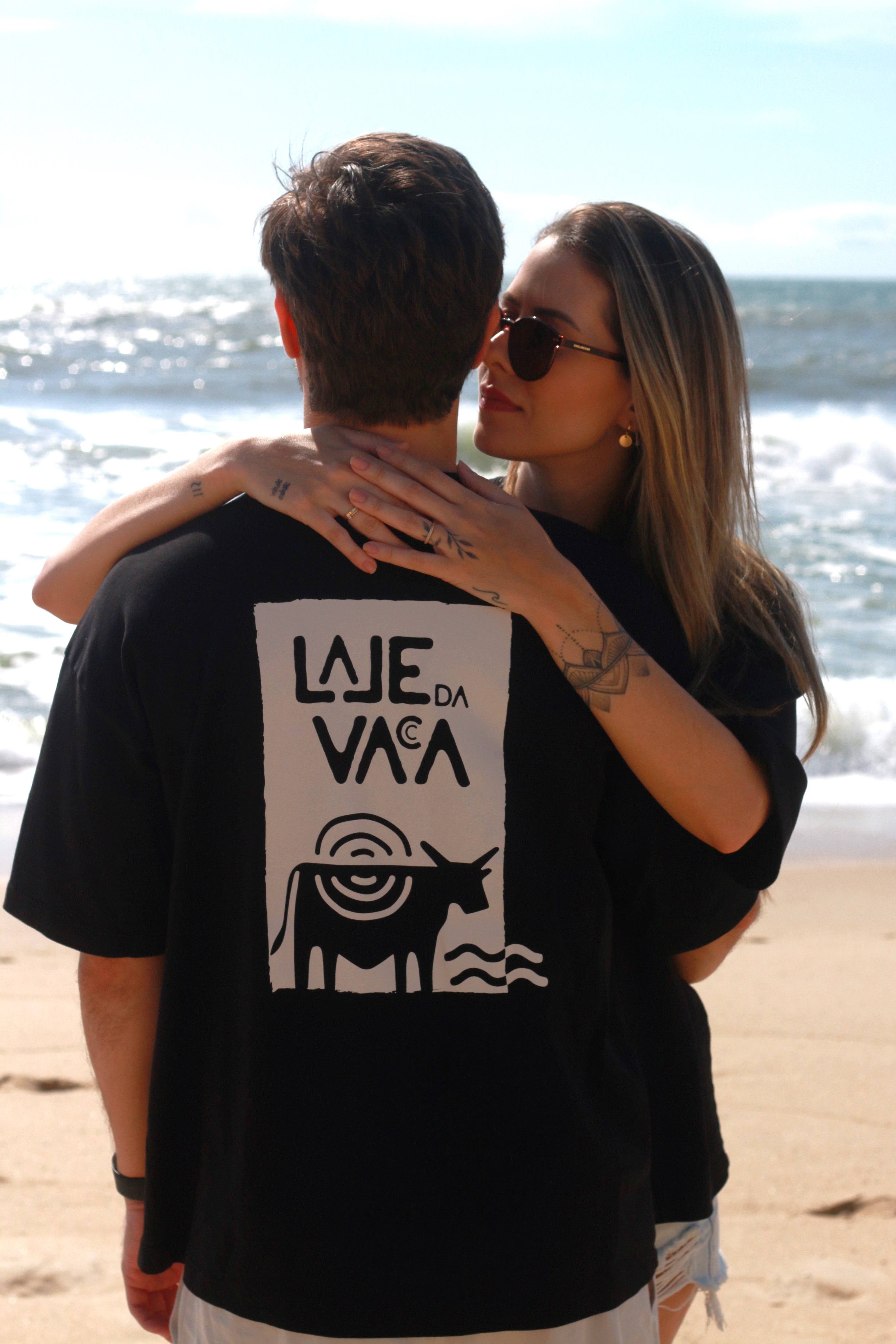 Casal na praia com a estampa Raízes Icon nas costas da camiseta preta.